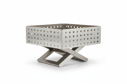 A portable, fold-flat stainless steel fire pit with a modern, minimalist design. The fire pit is made of brushed stainless steel panels that fold together, forming a shallow square bowl with perforated sides for airflow. It sits on a sturdy X-shaped metal base that elevates it off the ground. The panels have evenly spaced round ventilation holes and visible metal fasteners at the corners. The fire pit is shown assembled, centered on a clean white background, photographed at a slight front-angle, with sharp 